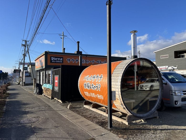 青空の下、D'STYLEの看板の前に置かれたオレンジ色のバレルサウナ。丸い木口とガラス扉、屋根から煙突が立つ