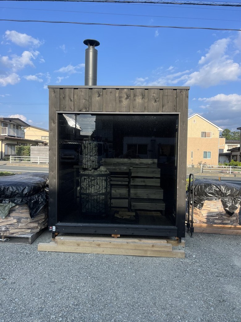 青空の下、黒い板張りのサウナ小屋を正面から。大きなガラス面ごしに石を積んだヒーターと段状のベンチが見える