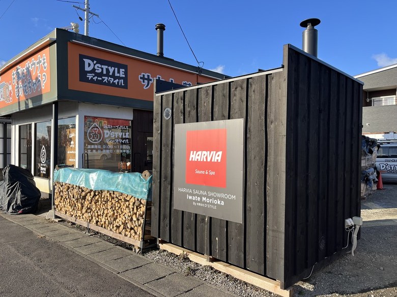 黒い板張りのサウナ小屋。壁にHARVIA SAUNA SHOWROOM Iwate Morioka の赤いパネル。左に薪棚と店舗