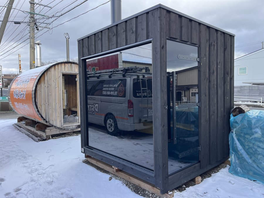雪の店先に並ぶ黒いキューブサウナとバレルサウナ