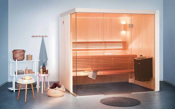 青みがかった灰色の壁の部屋に置かれた、ガラス張りの木のサウナ室。手前に白い棚とピンクのスツール、籠が並ぶ