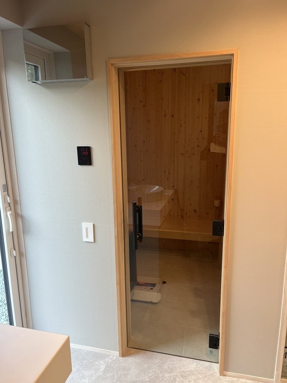 当社施工:脱衣室からサウナ室のガラス扉を見たところ。壁に黒いコントローラーと照明スイッチ